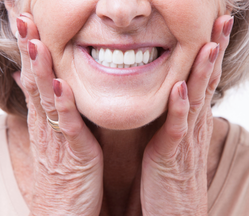 Close-up of a smiling older woman cupping her cheeks with both hands, showing white teeth.