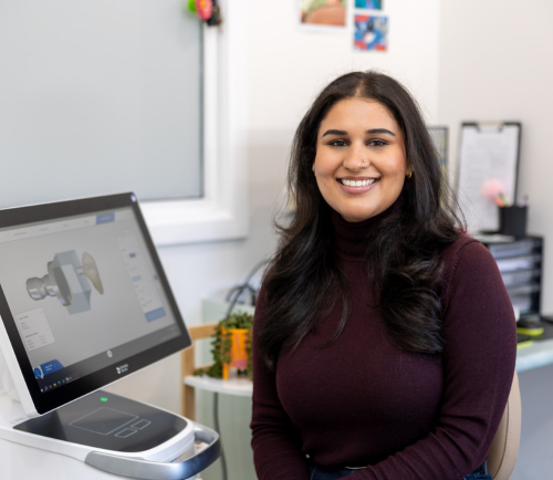 Smiling woman sits at a desk next to a computer showing a 3D model on the screen in a modern design office.