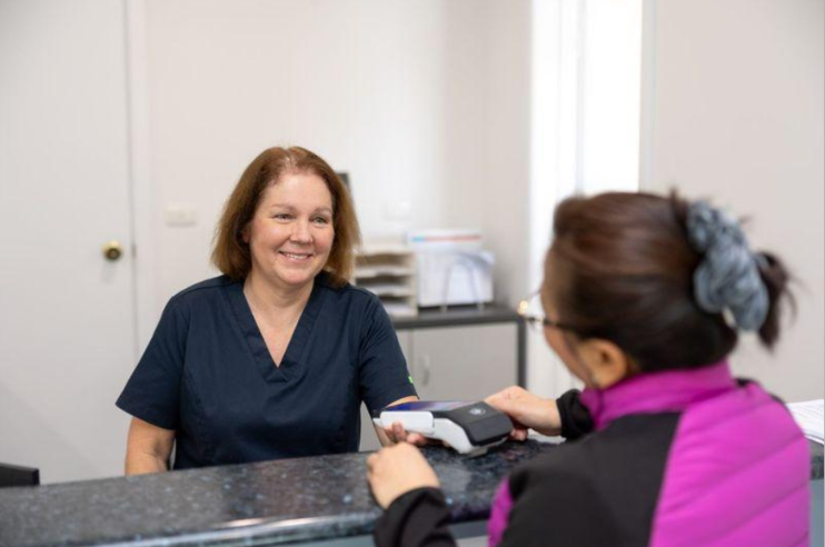 Smiling healthcare worker in navy scrubs assists a patient at a clinic front desk with a card payment terminal on the counter.